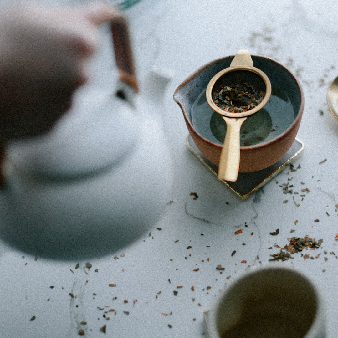 Tea-making process with a ceramic teapot and strainer on a light surface.