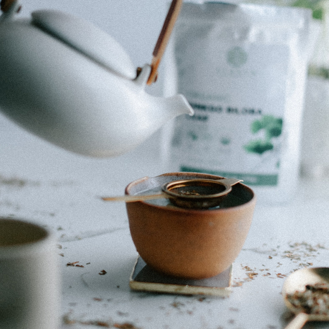 Tea being poured from a white teapot into a brown ceramic cup on a light surface.