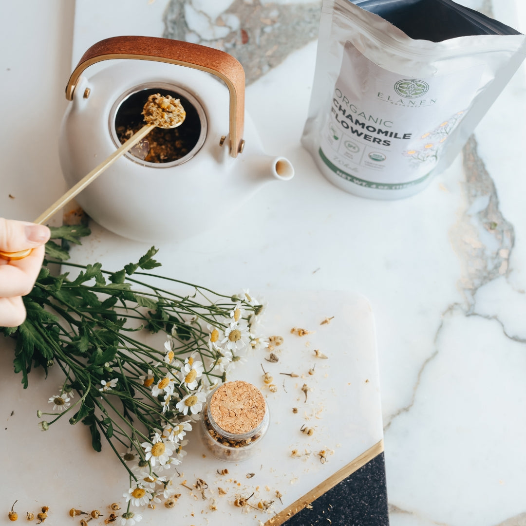 Tea-making setup with a teapot, chamomile flowers, and packaging on a marble surface.