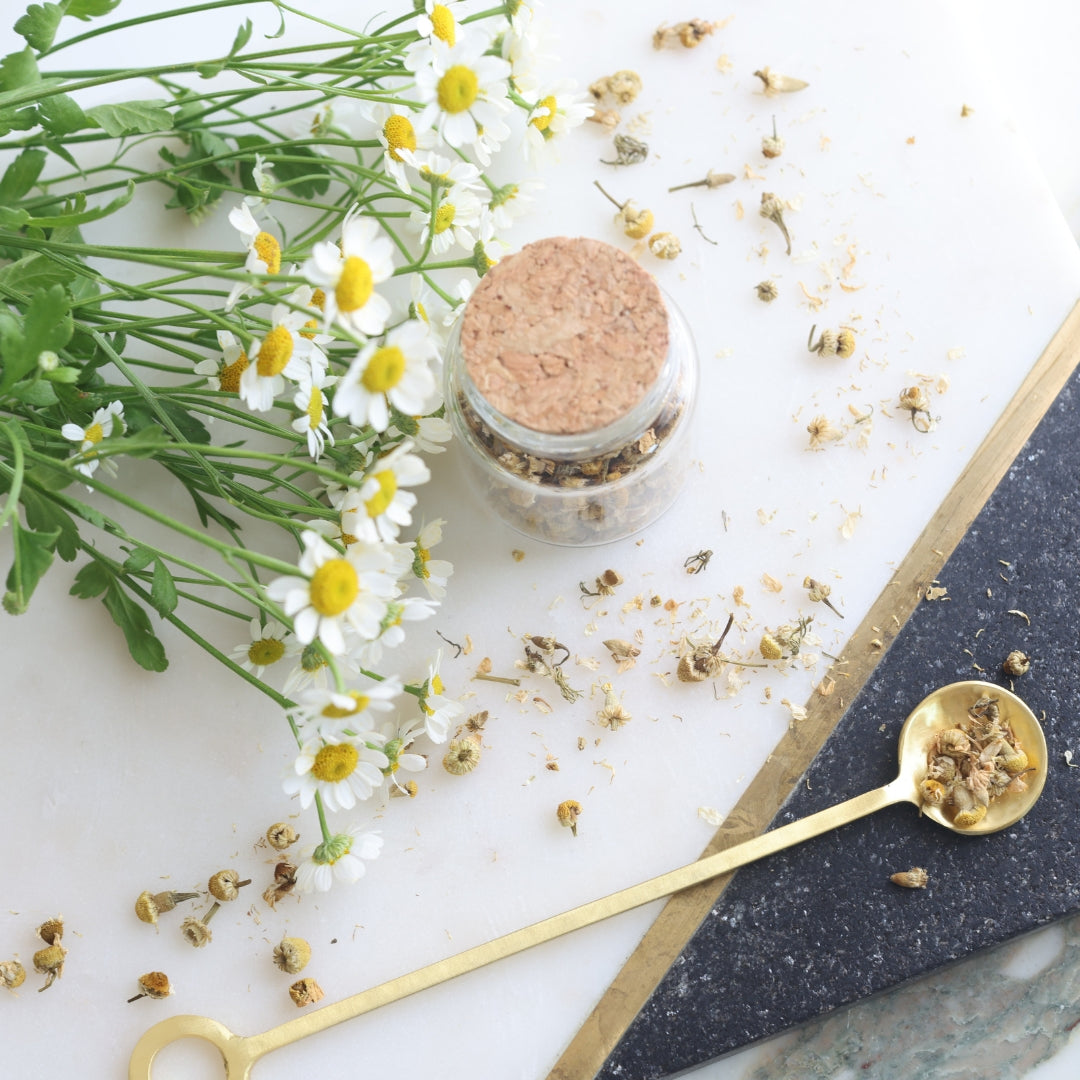 Small glass jar with cork lid, gold spoon with dried chamomile, and flowers on a white surface