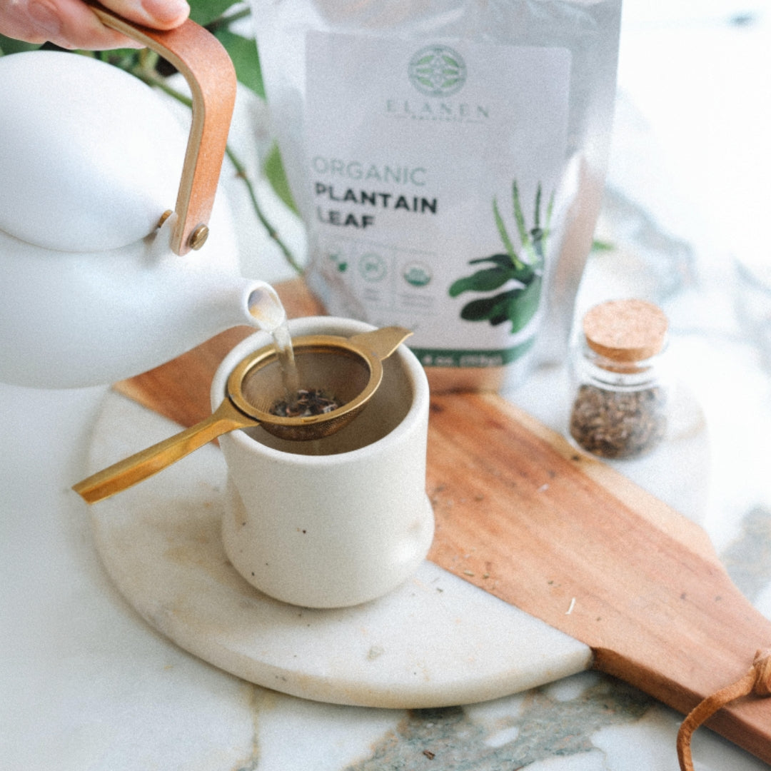 Tea being poured into a cup with a package of organic plantain leaf tea in the background.