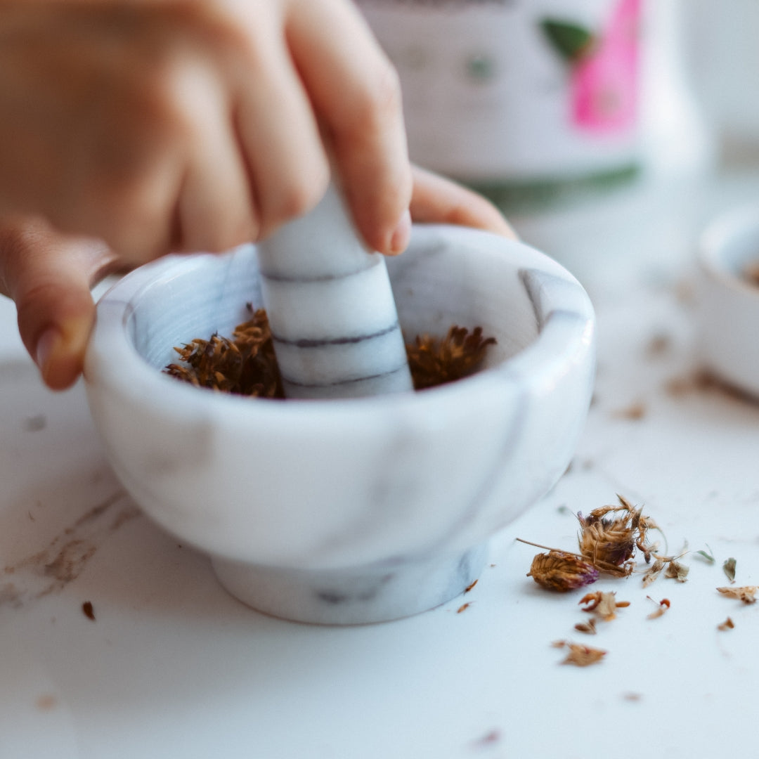 Person using a marble pestle to crush red clover in a mortar on a light surface.