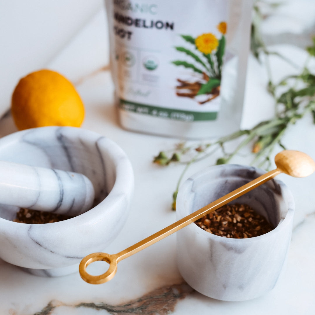 Marble mortar and pestle with gold handle on a marble surface with a container of dandelion root in the background.
