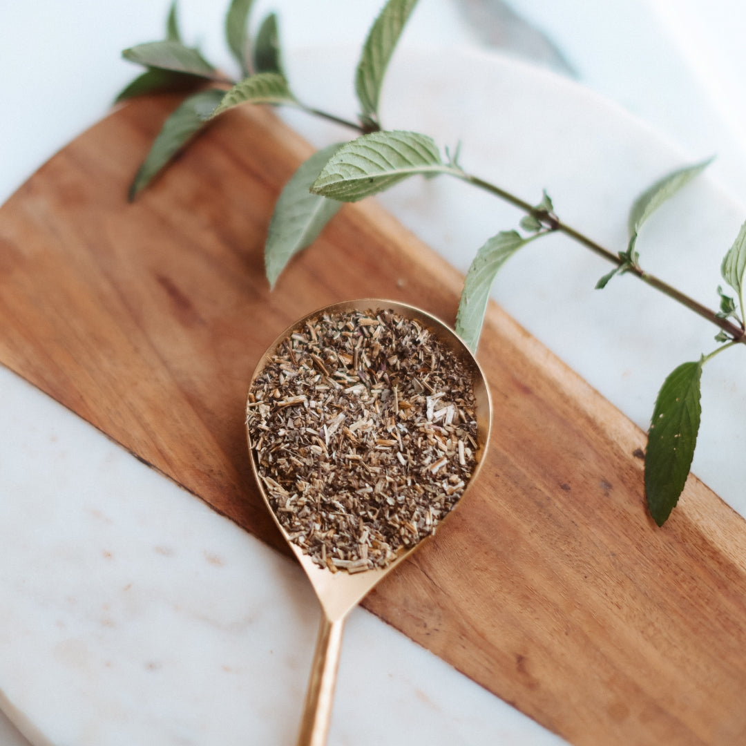 Gold spoon filled with dried peppermint leaf on a wooden cutting board with green leaves.