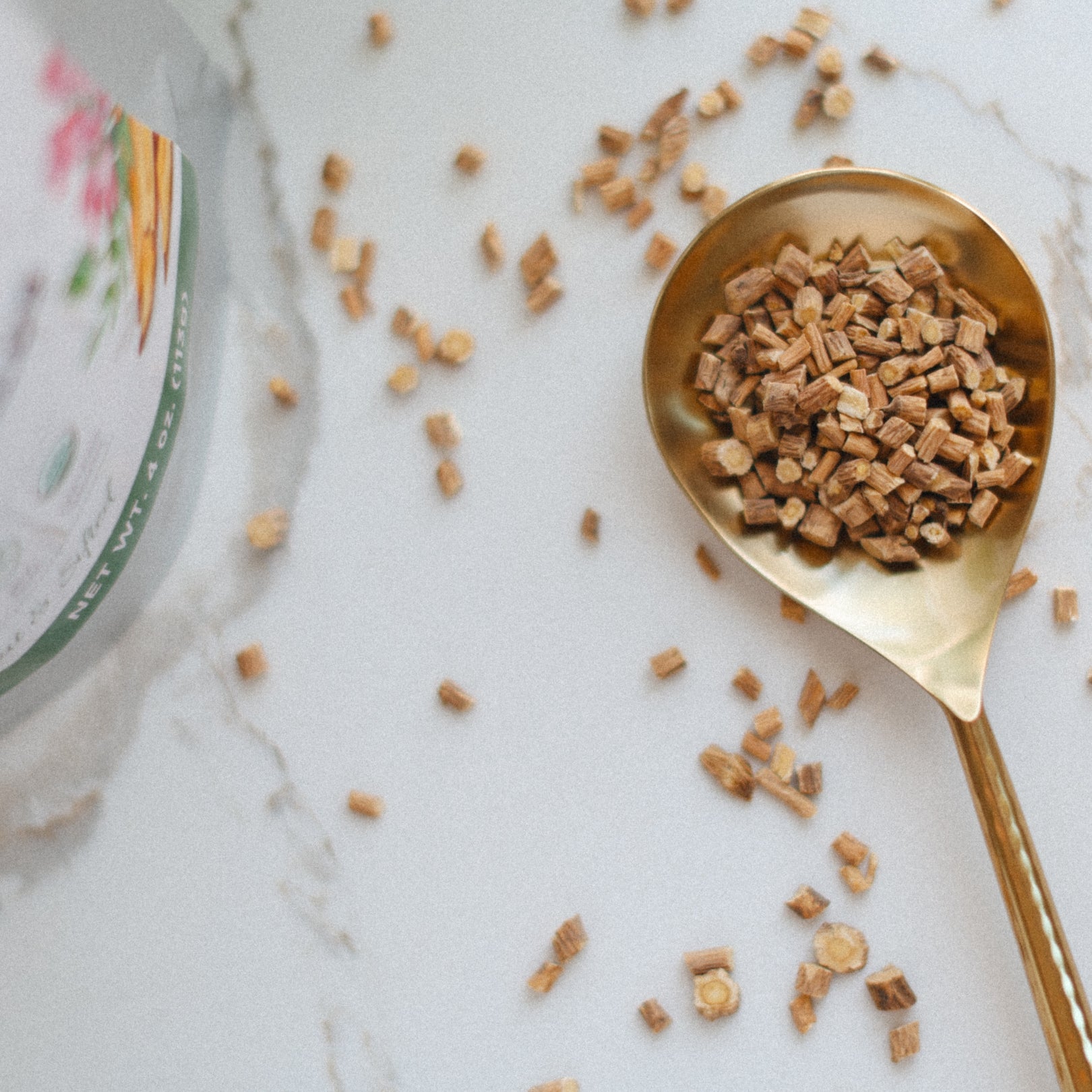 Gold spoon filled with astragalus root pieces on a marble surface with a container of sugar in the background.