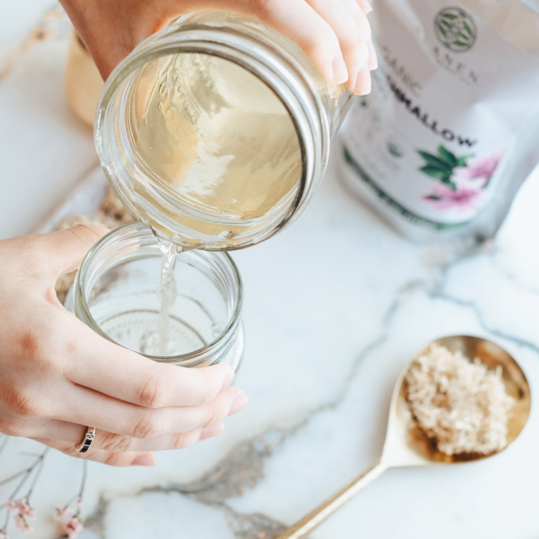 Person pouring a liquid from one jar into another on a marble surface with a spoon and product packaging.