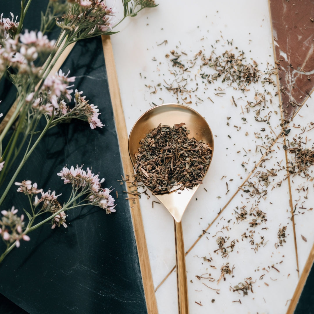 Gold spoon filled with dried valerian root on a white surface with flowers and leaves.