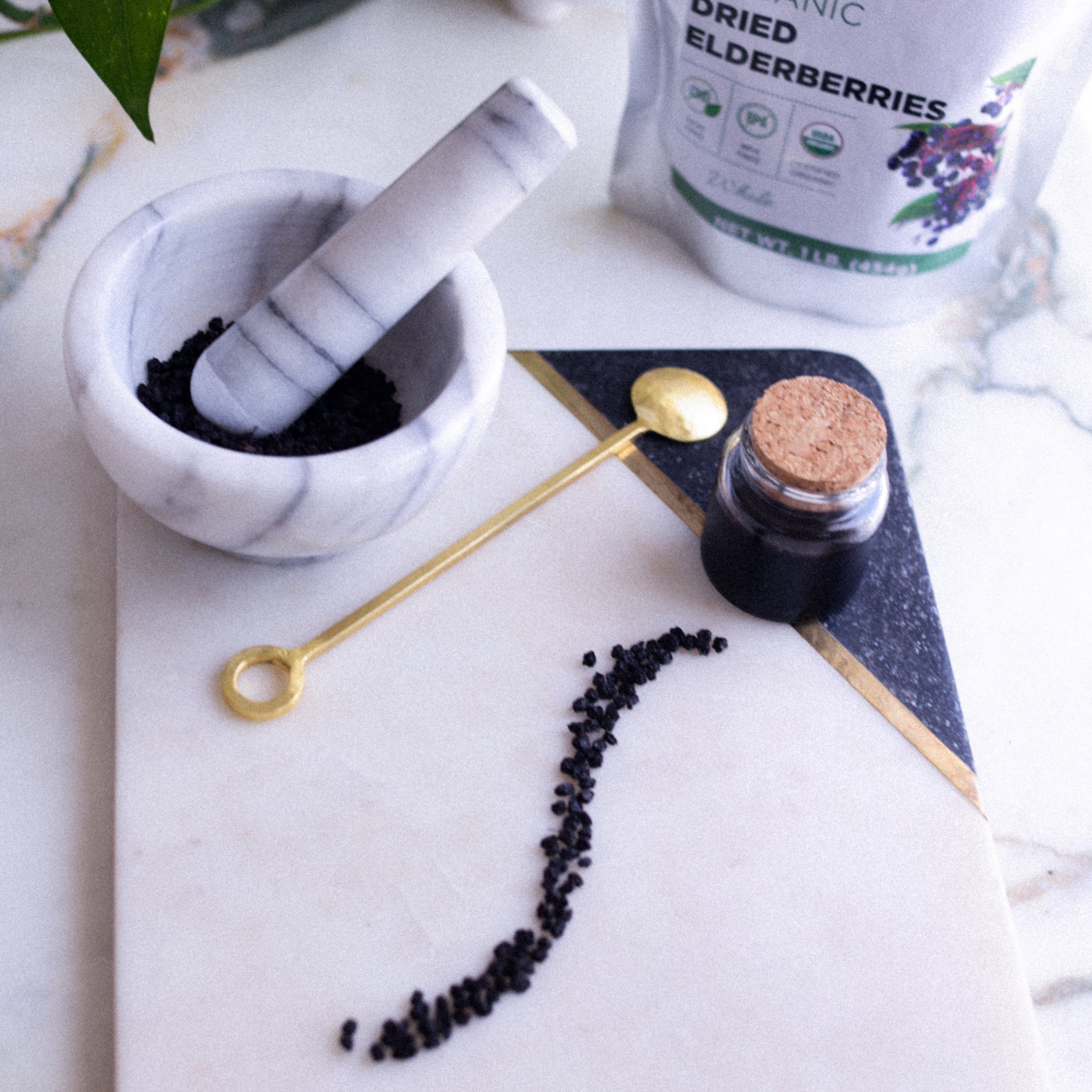 Marble mortar and pestle with black elderberries, jar of elderberry syrup, and spoon on a marble surface.