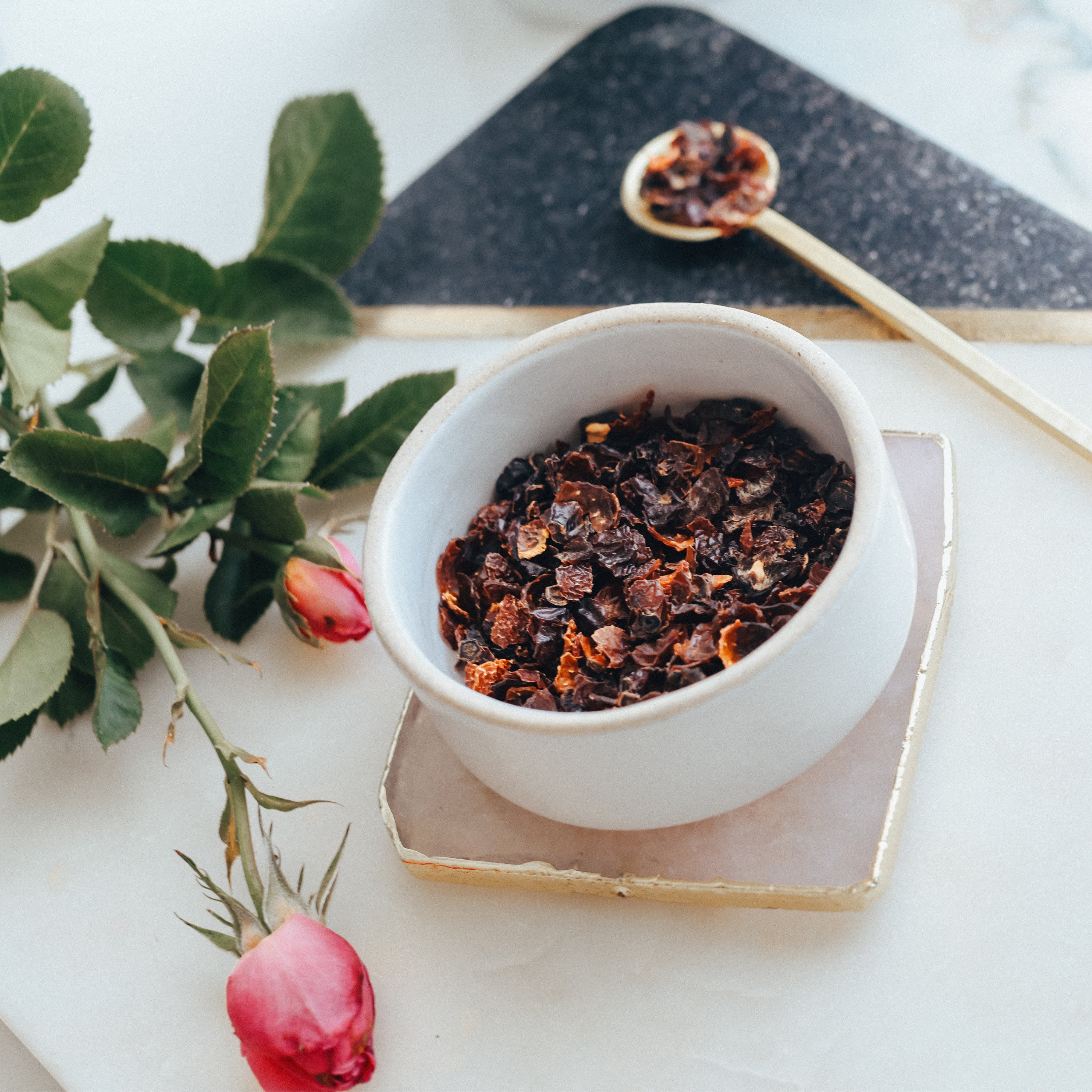 White bowl with dried rosehips on a white surface with green leaves and pink roses.