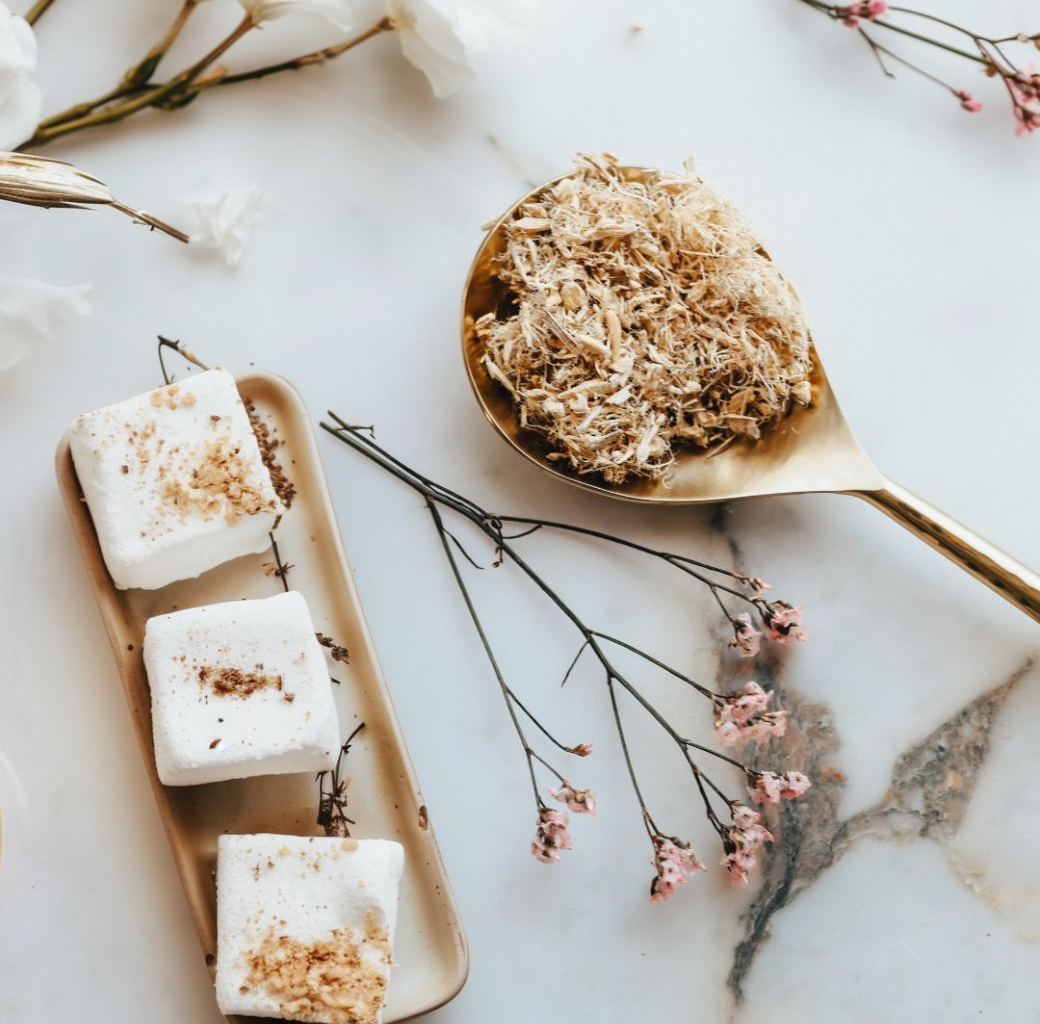 White marshmallows with brown toppings on a beige plate, accompanied by a gold spoon filled with more marshmallow root, all on a marble surface with floral elements.