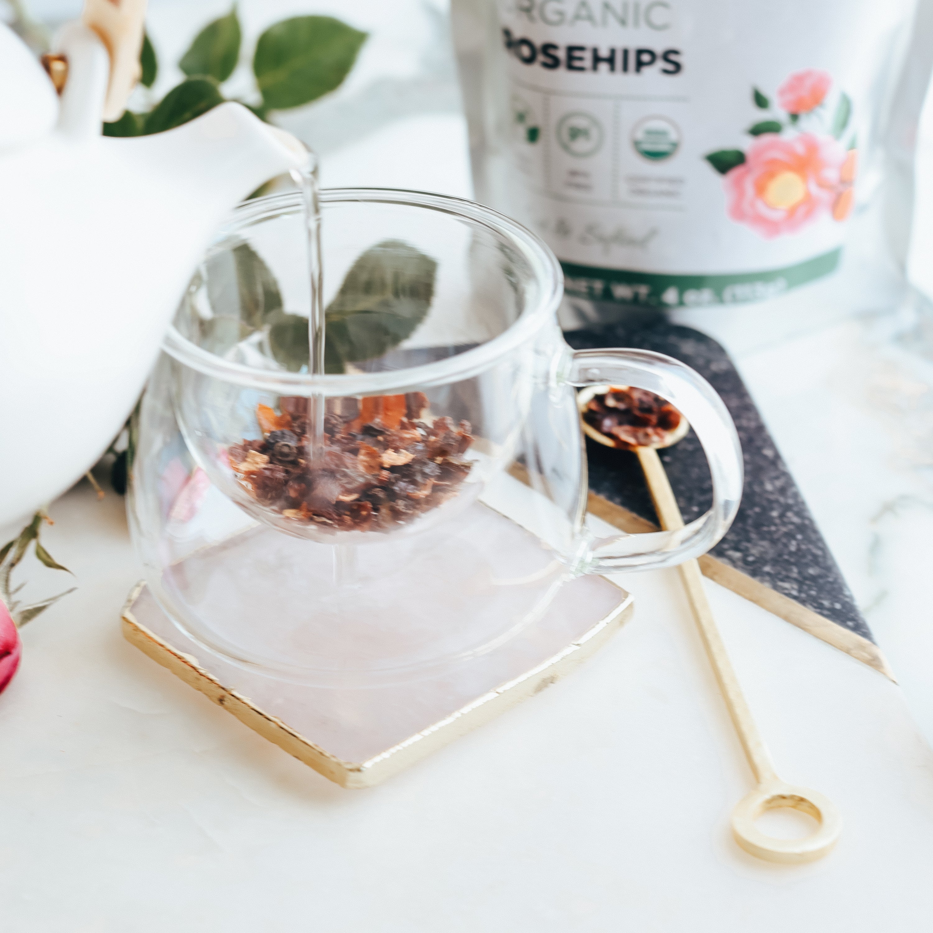 Glass teacup with rosehips, gold spoon, and container labeled 'Organic Rosehips' on a white surface.