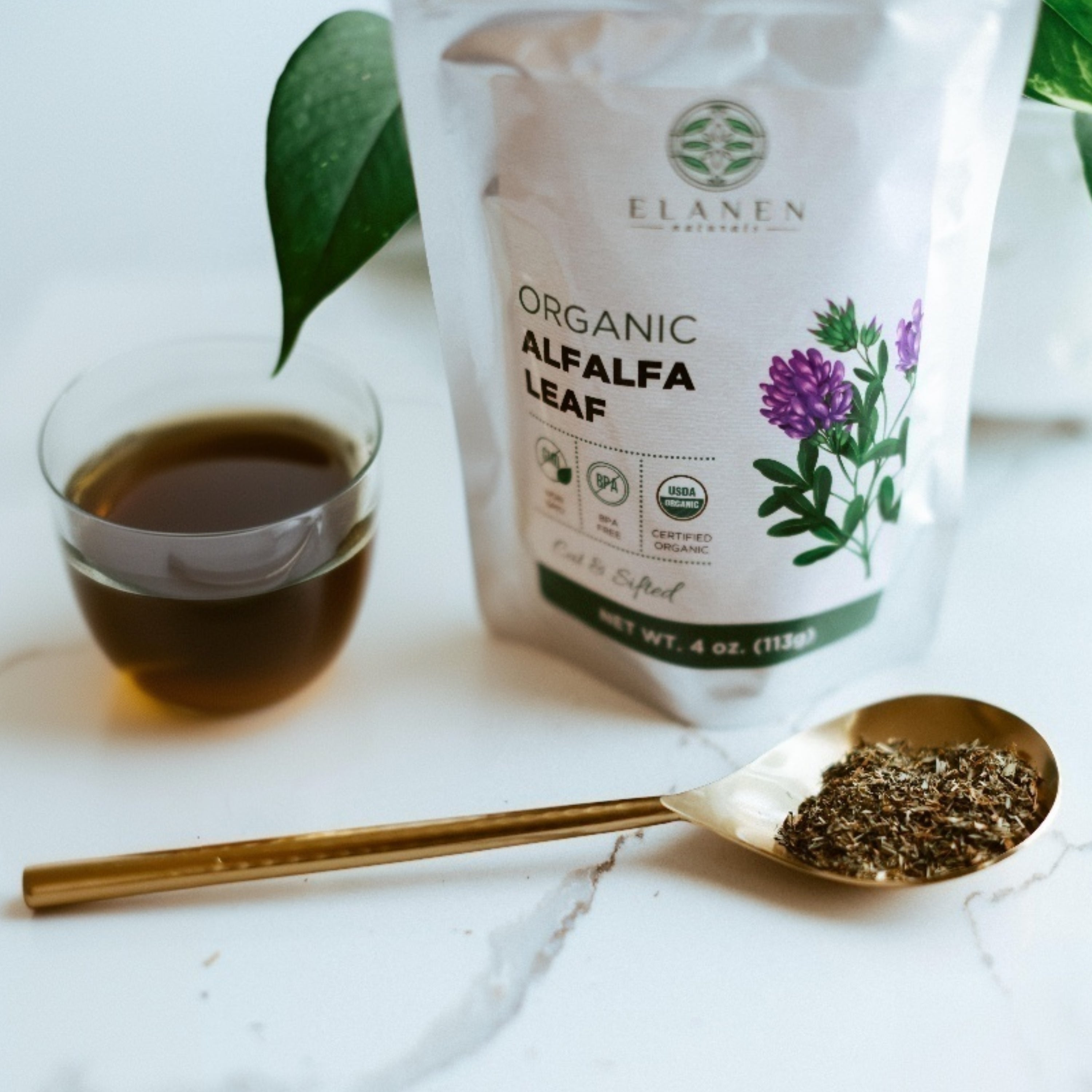Elanen Organic Alfalfa Leaf packaging with a spoonful of alfalfa leaves and a cup of tea on a marble surface.