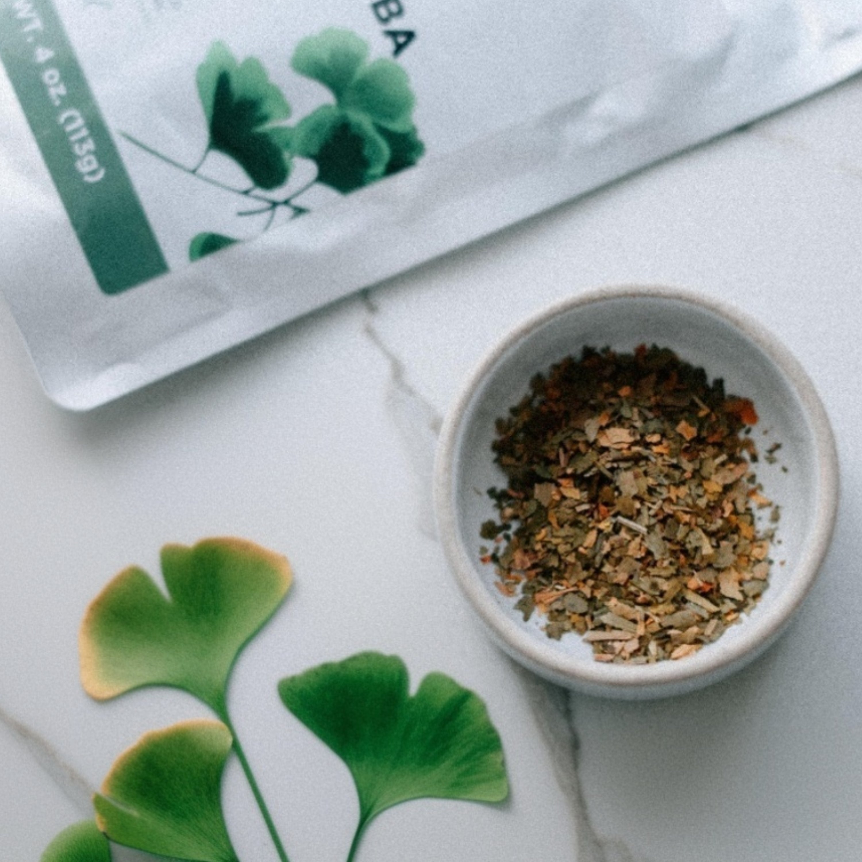Small bowl of dried herbs on a white surface with green leaves and a package in the background.