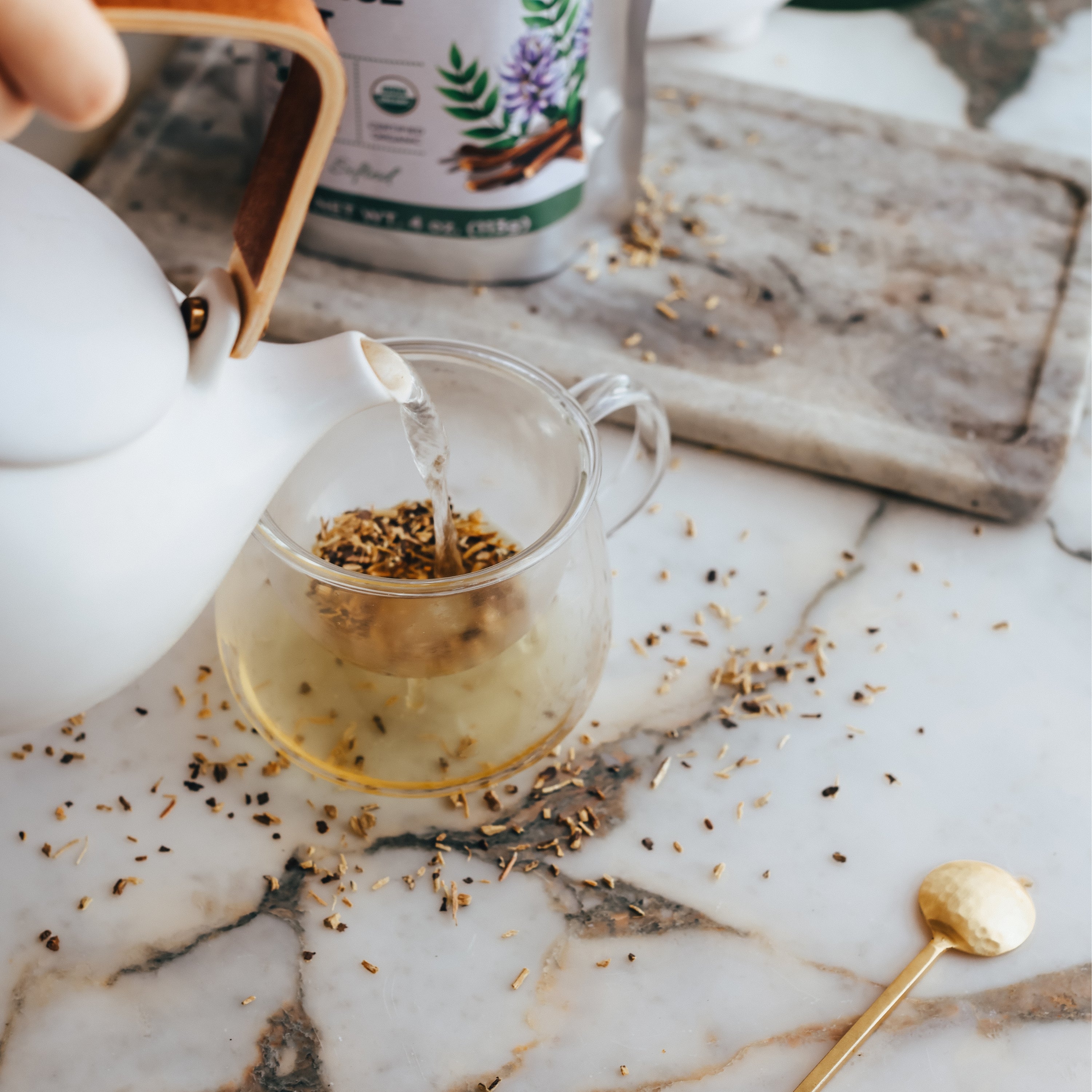 Tea being poured into a glass mug with licorice root on a marble surface with a tea bag in the background.