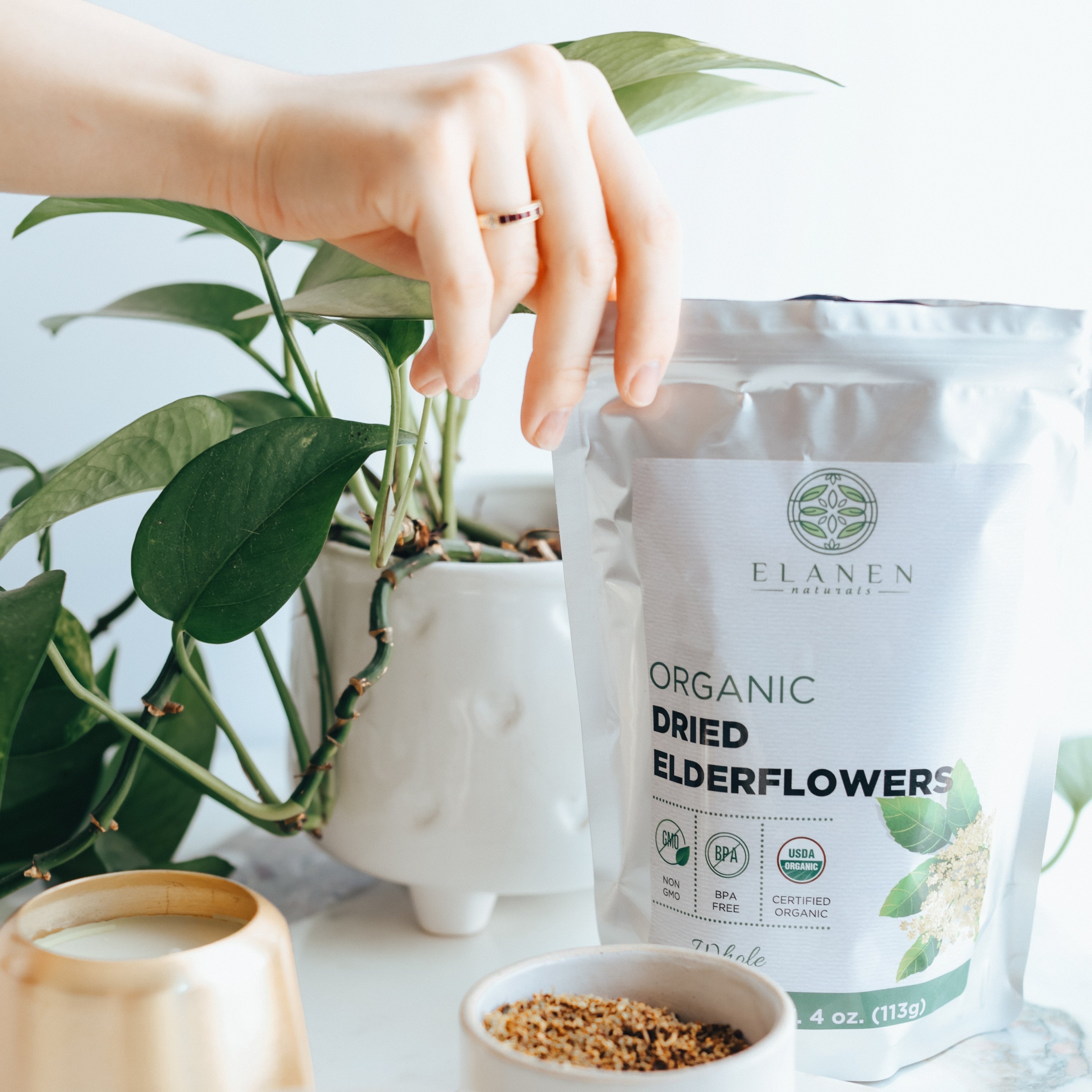 Hand holding a package of Elanen Organic Dried Elderflowers with a plant and cup in the background.