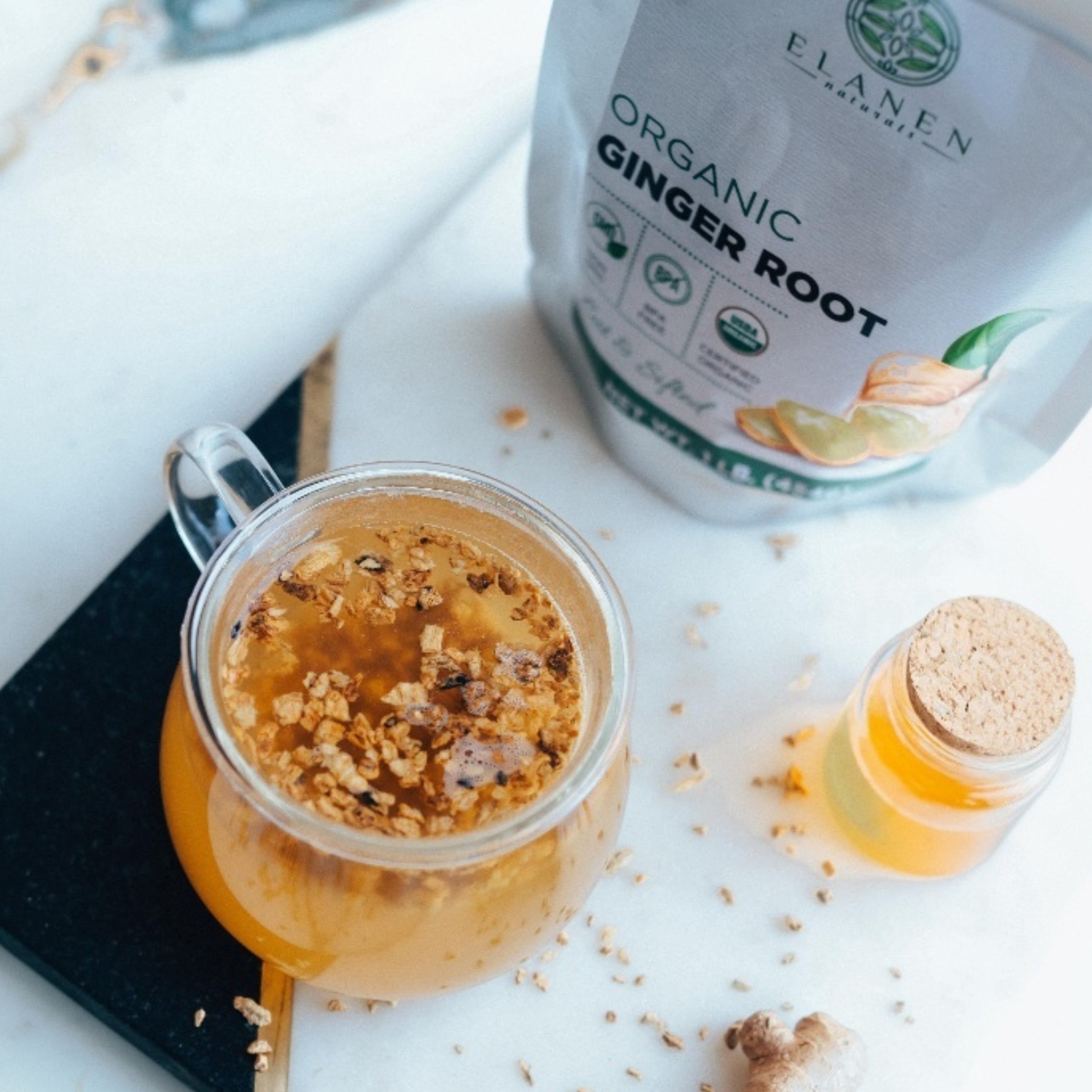 Glass mug with ginger tea, ginger root, and a bottle of honey on a white surface with Elanen Organic Ginger Root packaging.