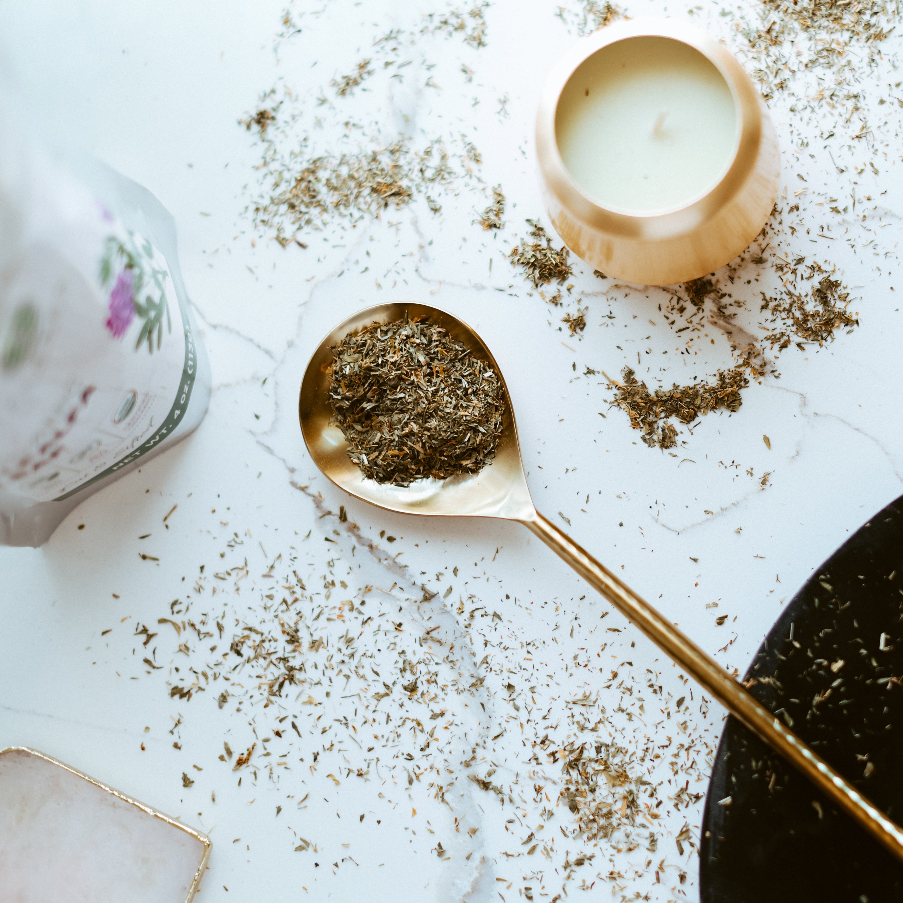 Gold spoon with dried herbs on a light surface with a candle and tea.