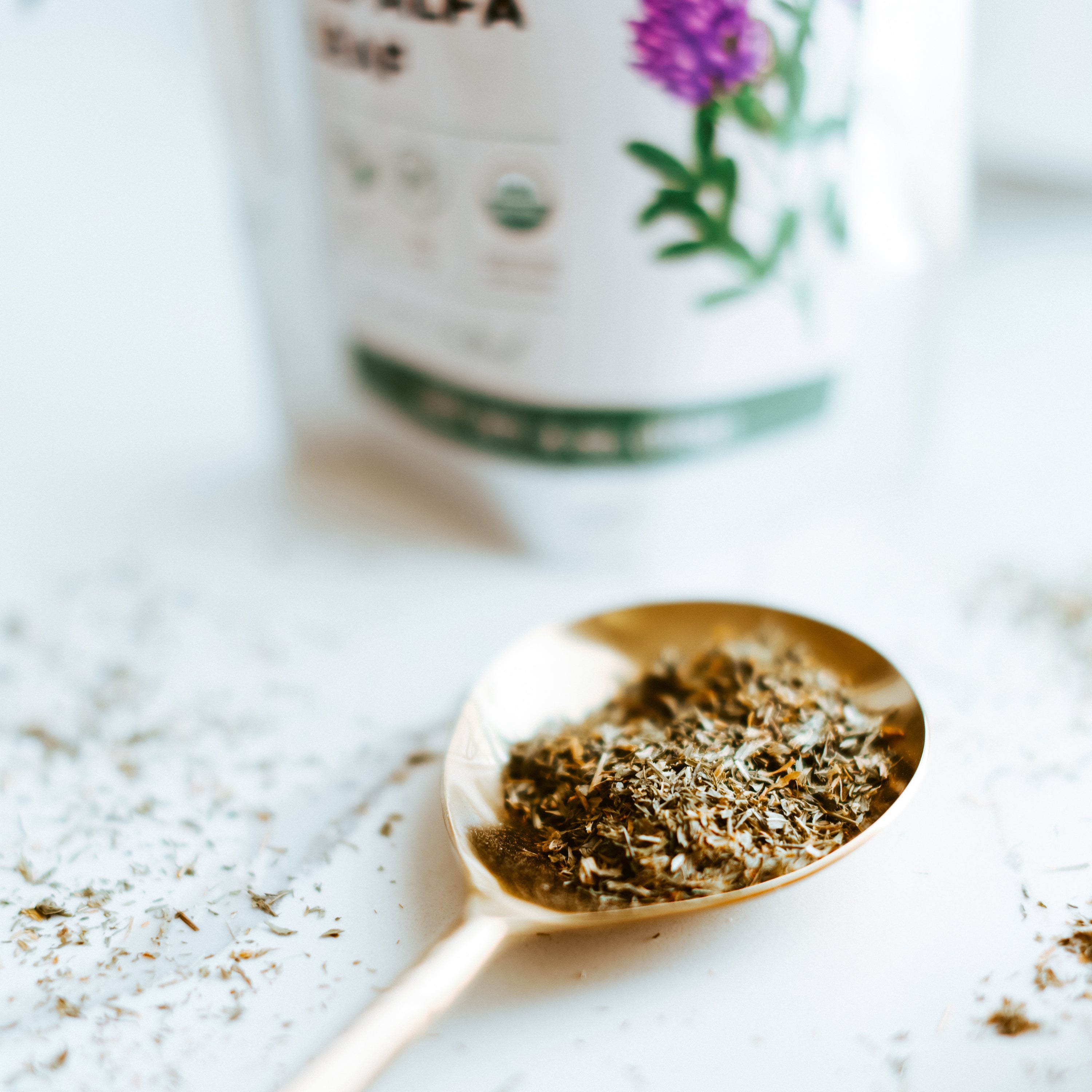 Gold spoon filled with dried herbs on a light surface with a blurred container in the background.