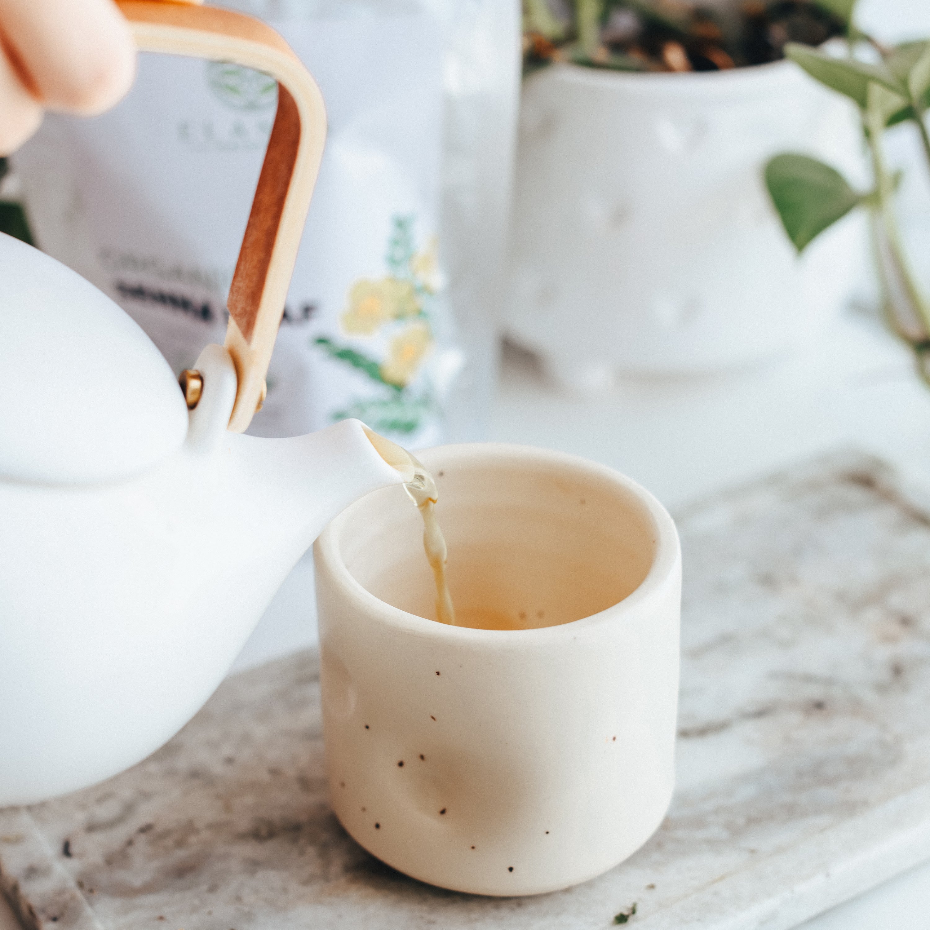 Tea being poured from a white teapot into a beige ceramic cup on a marble surface.