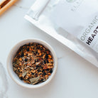 Tea leaves in a white bowl with an open package of Elanen Organic Heart Tea in the background.