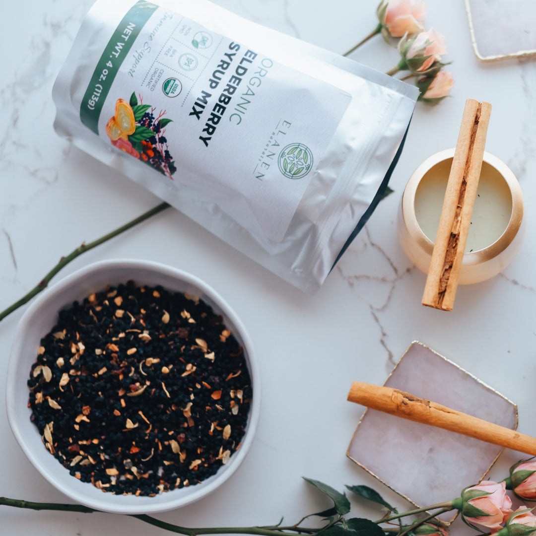 Tea packaging, bowl of elderberry syrup mix, and cinnamon sticks on a marble surface with flowers.