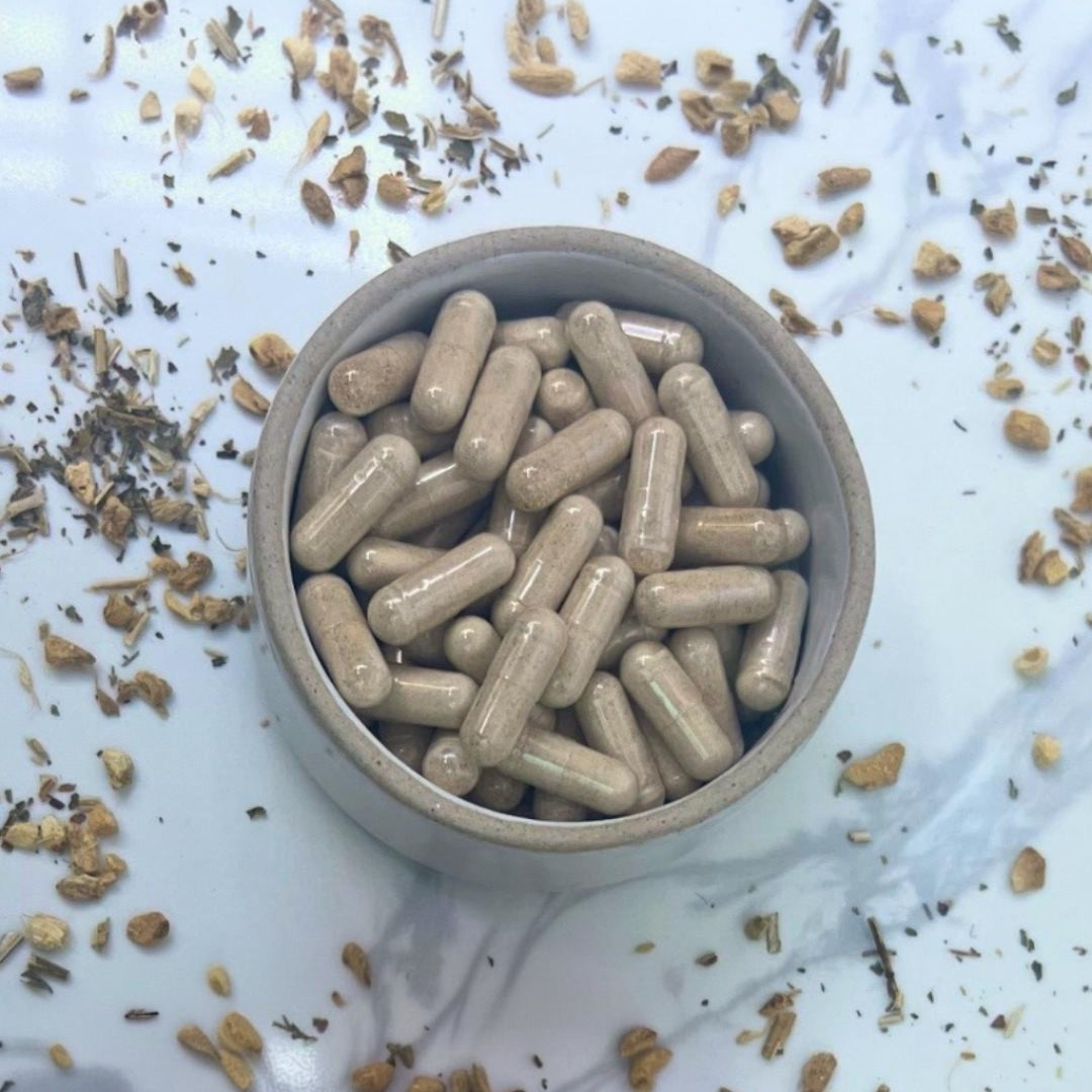Small bowl filled with beige capsules on a white surface with dried herbs.