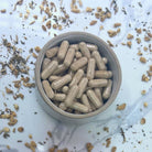 Small bowl filled with beige capsules on a white surface with dried herbs.