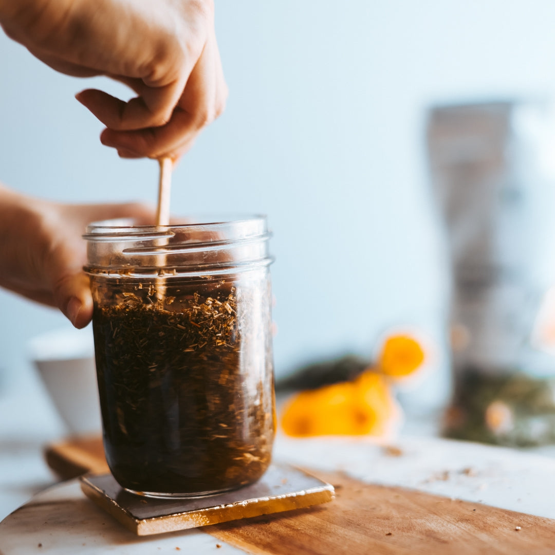 Person stirring a jar of tea with a spoon on a wooden coaster.