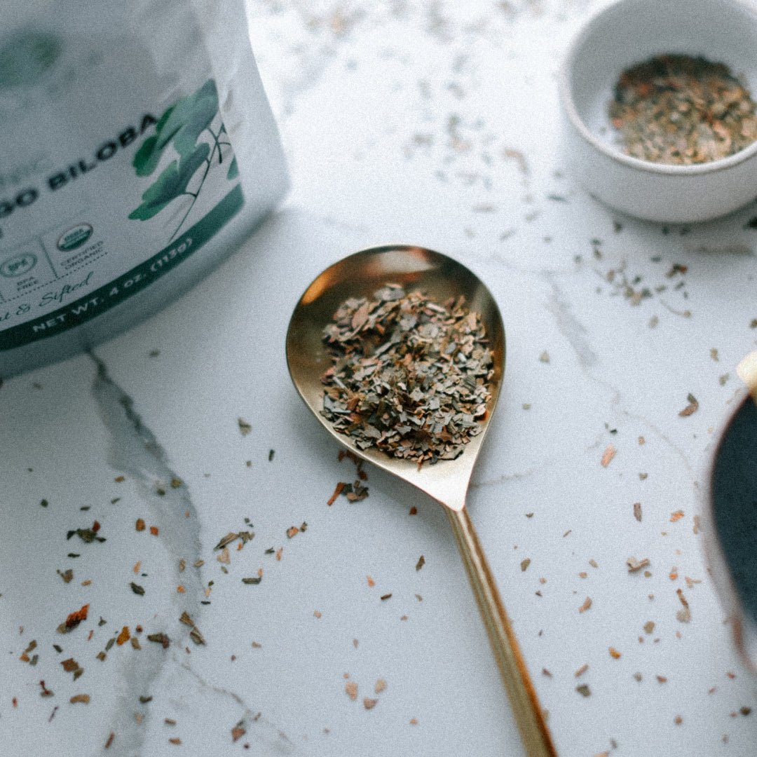 Gold spoon with dried herbs on a marble surface next to a bag of organic biloba leaf.