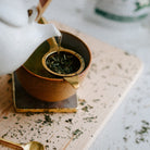 White teapot pouring hot water into a brown ceramic cup on a textured surface.