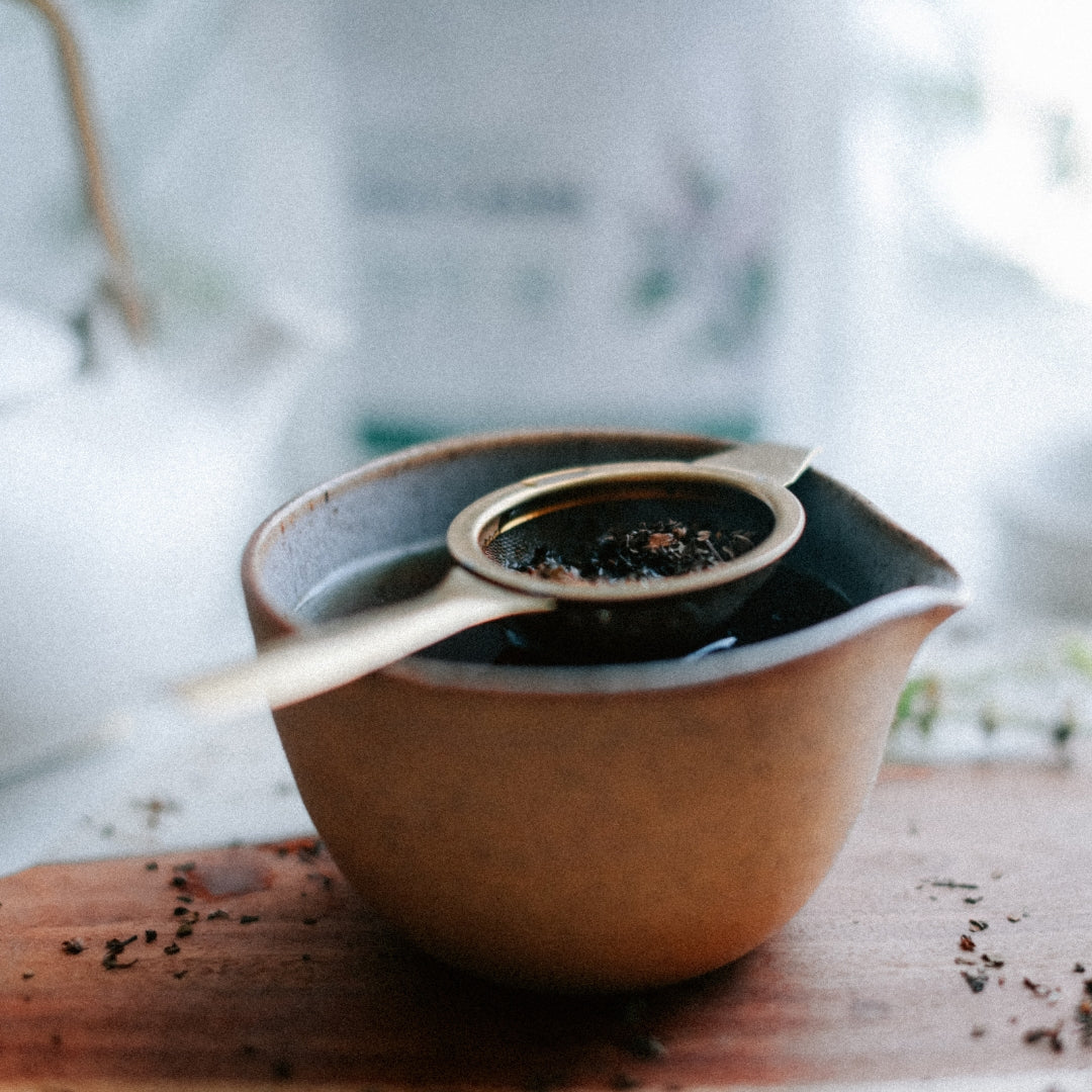 Ceramic tea strainer with loose leaf tea in a teapot on a wooden surface.