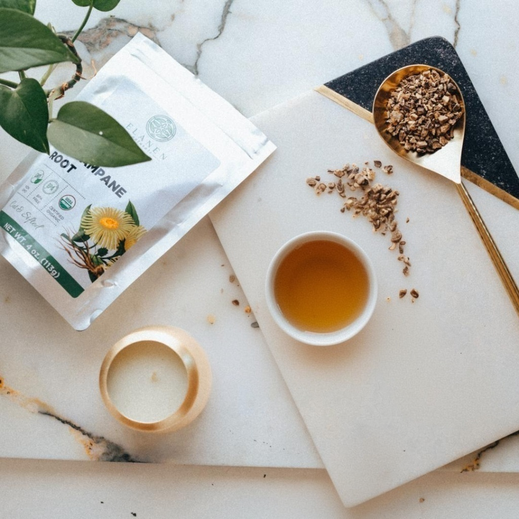Tea packaging, cup of tea, and spoon with elecampane root on a marble surface