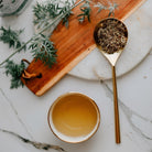 Tea cup, gold spoon with wormwood leaves, and wooden board on a marble surface