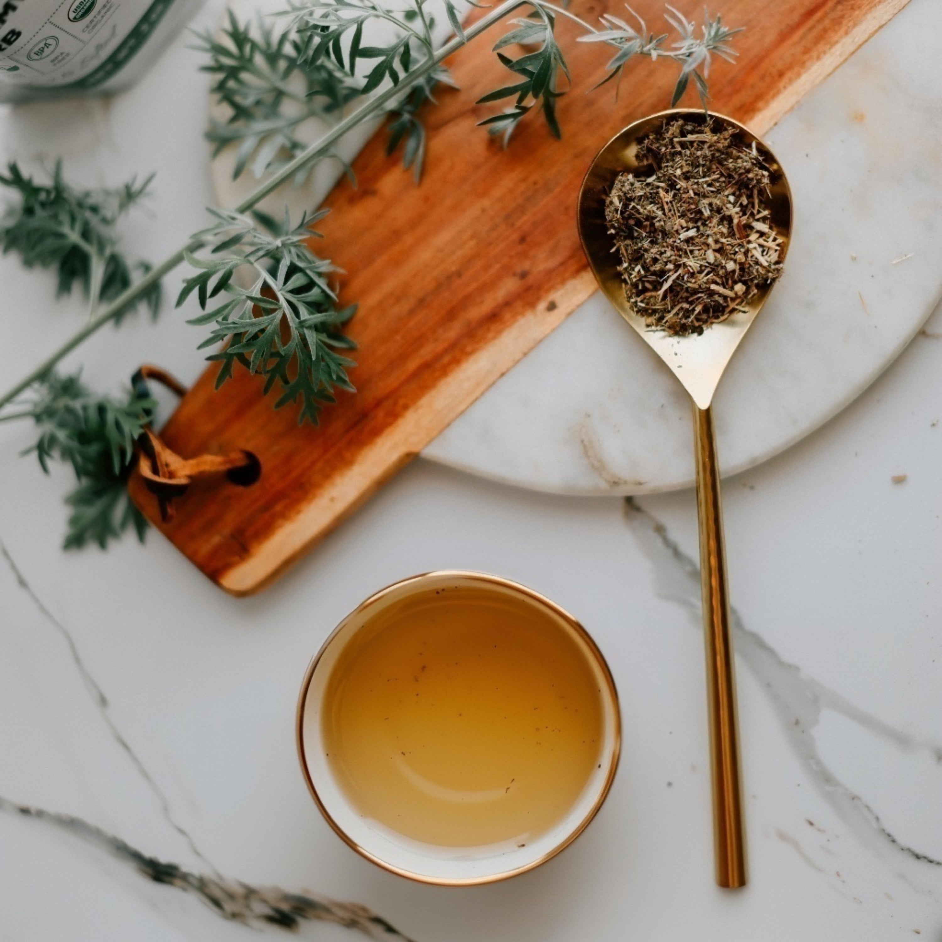 Tea cup, gold spoon with wormwood leaves, and wooden board on a marble surface