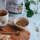 Tea packaging, cup of tea, and bowl of tea leaves on a table with a plant in the background.