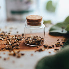 Small glass jar with cork lid on a wooden surface with dried herbs.