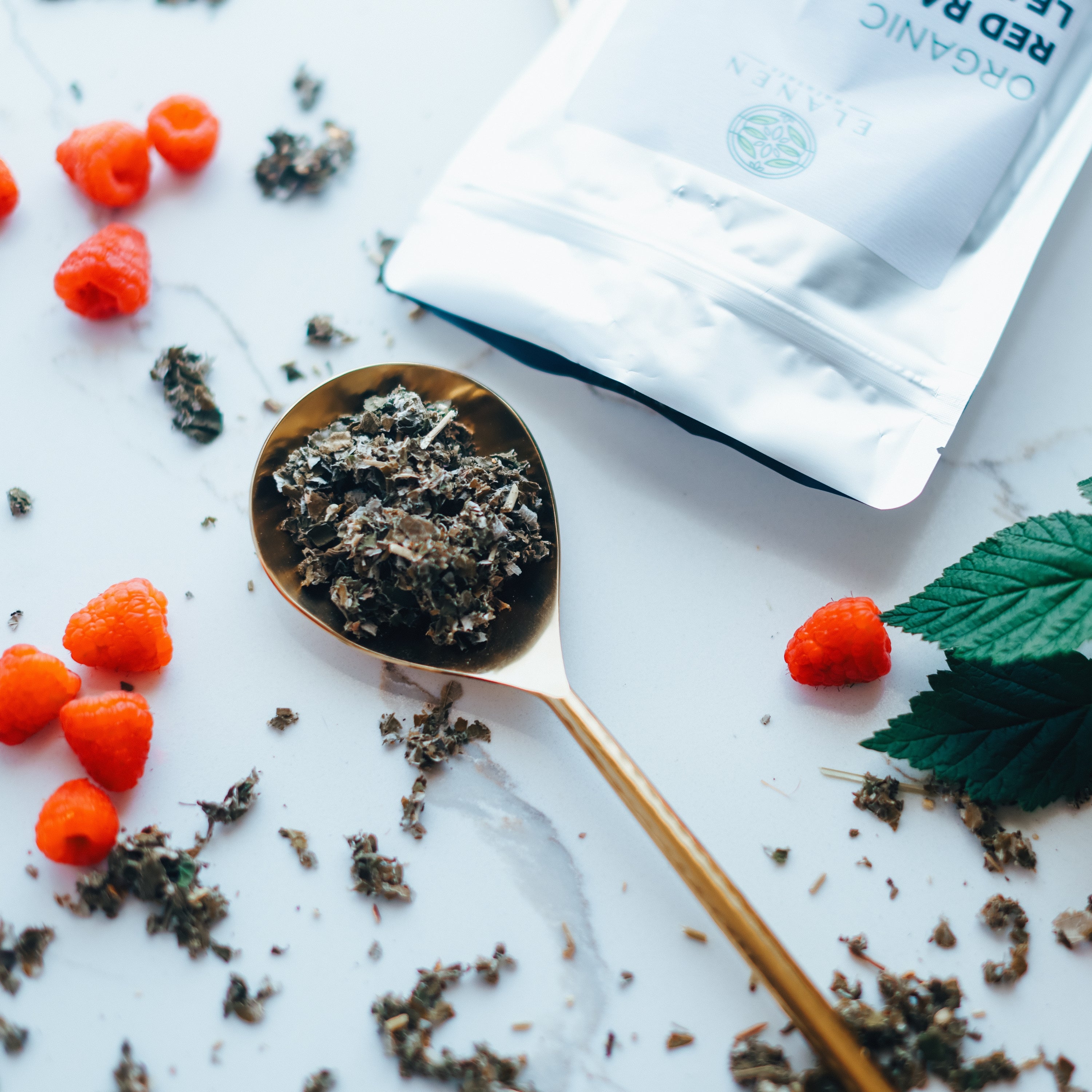 Tea leaves on a spoon with raspberries and a tea bag on a white surface