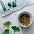 Small bowl of dried herbs on a white surface with green leaves and a package in the background.