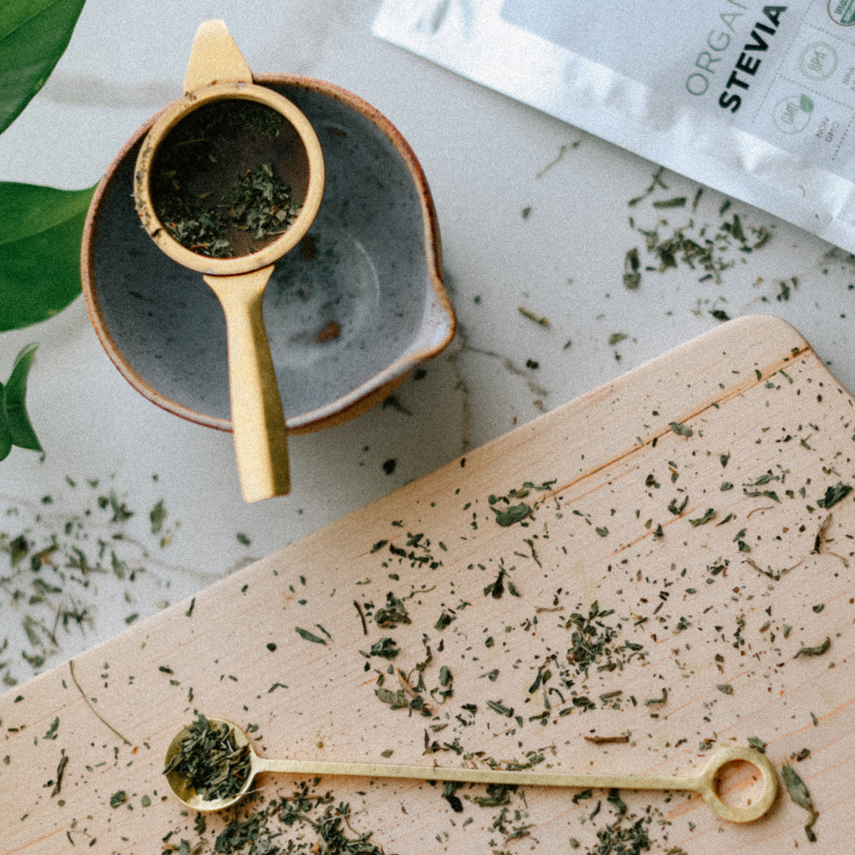 Stevia leaves in a small strainer over a bowl, with a wooden board and Stevia bag in the background.