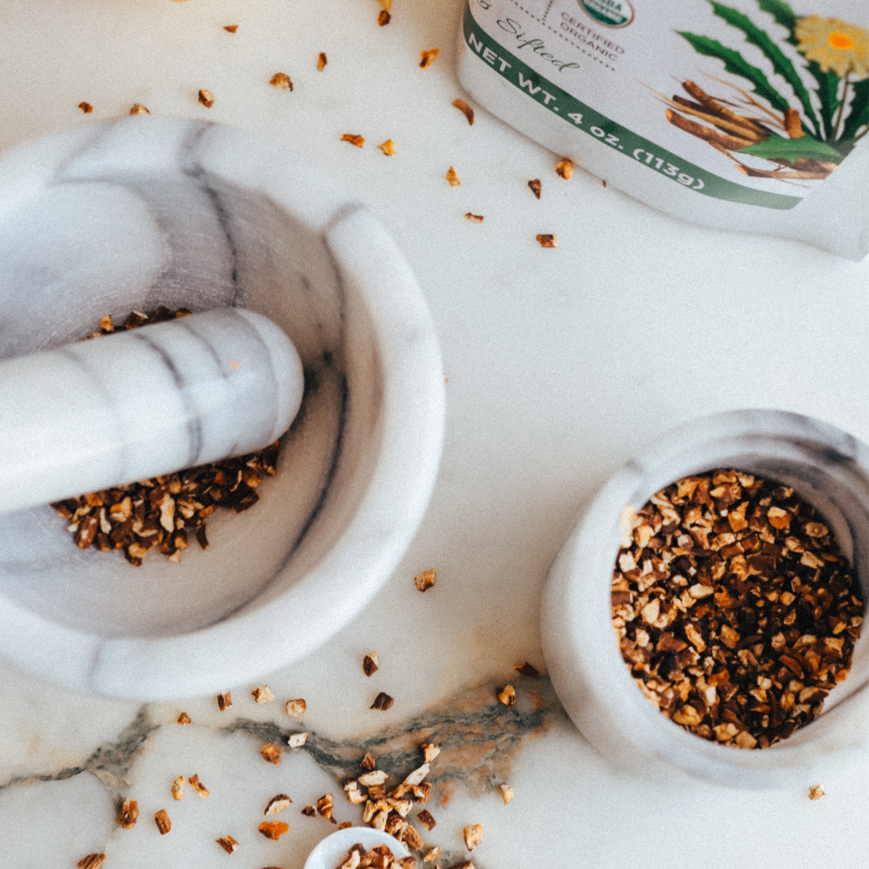 Mortar and pestle with ground dandelion root on a marble surface, next to a container of herbs.