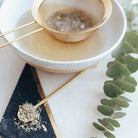 Tea being poured into a white ceramic cup with a gold spoon, surrounded by eucalyptus leaves on a light surface.