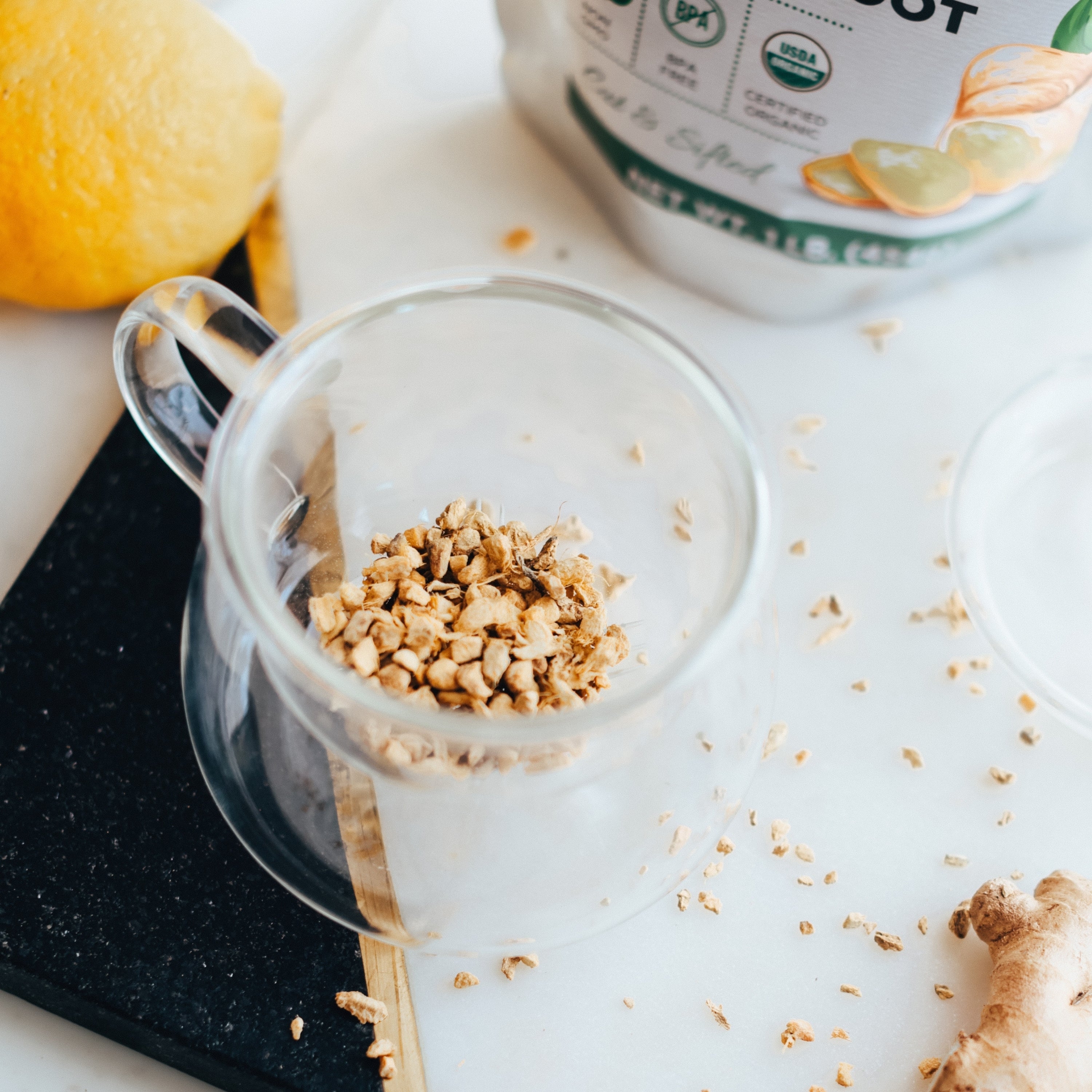 Clear glass mug with crushed ginger on a white surface with a lemon and container in the background.