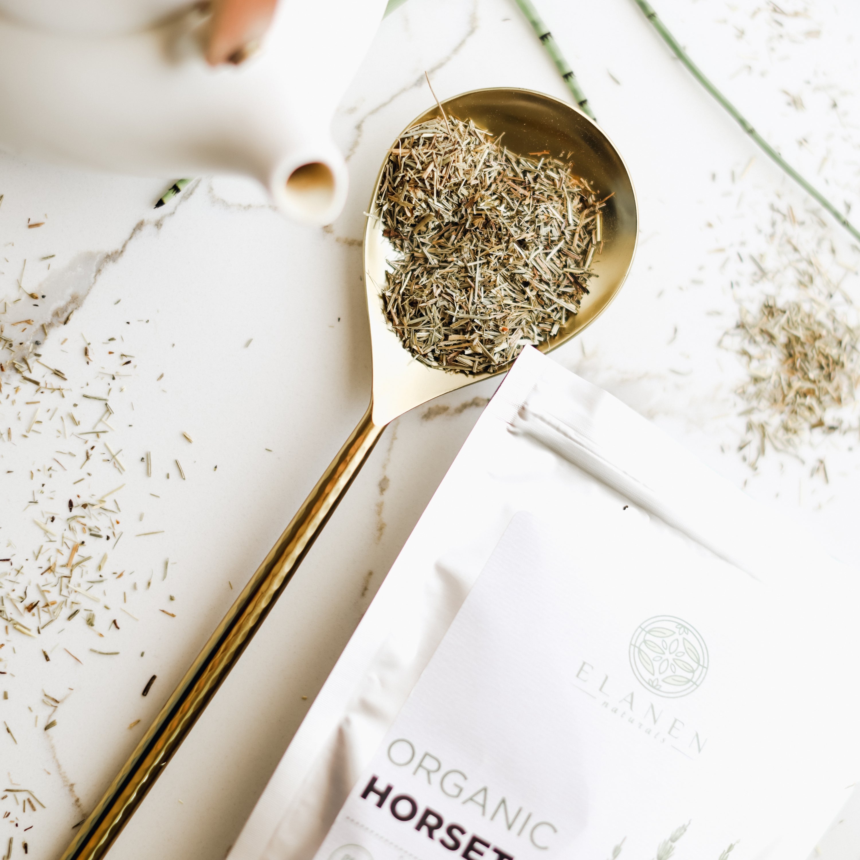 Gold spoon filled with dried herbs on a white surface with a package labeled 'Organic Horsetail' in the background.