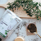 Spearmint leaves on a wooden board with a package of Flamen Organic Spearmint Leaf and a spoonful of spearmint leaves on a white surface.