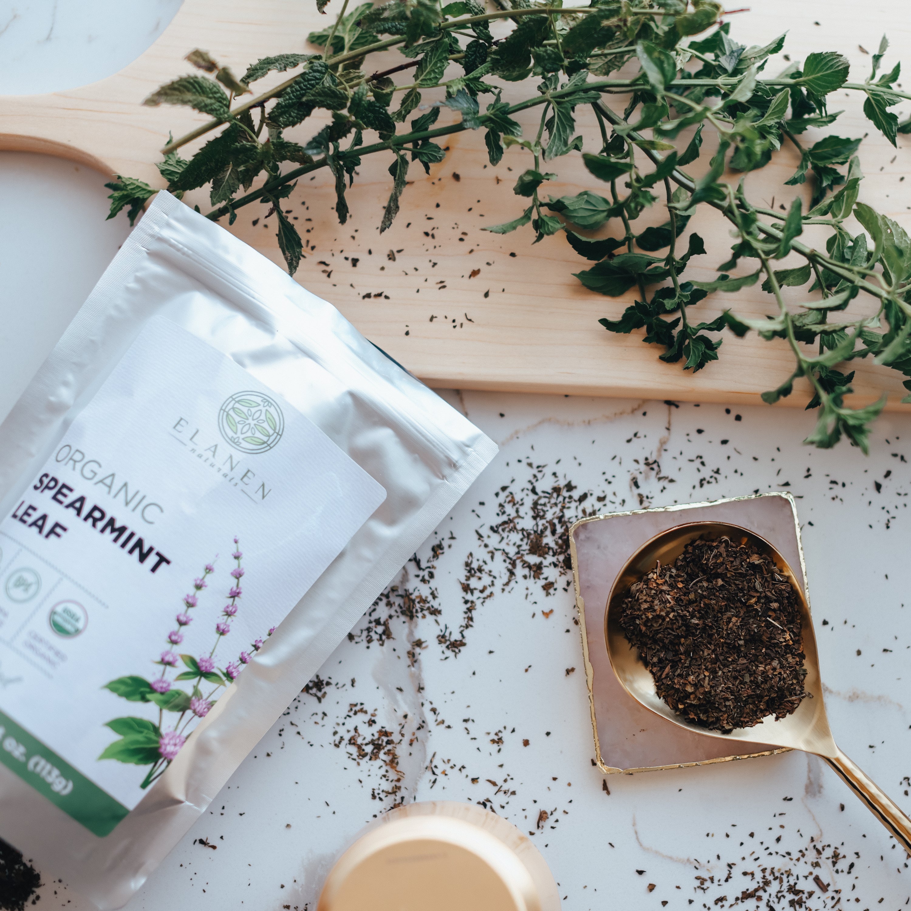 Spearmint leaves on a wooden board with a package of Flamen Organic Spearmint Leaf and a spoonful of spearmint leaves on a white surface.