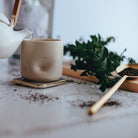 Tea being poured into a beige cup with a teapot, spoon, and leaves on a light surface.